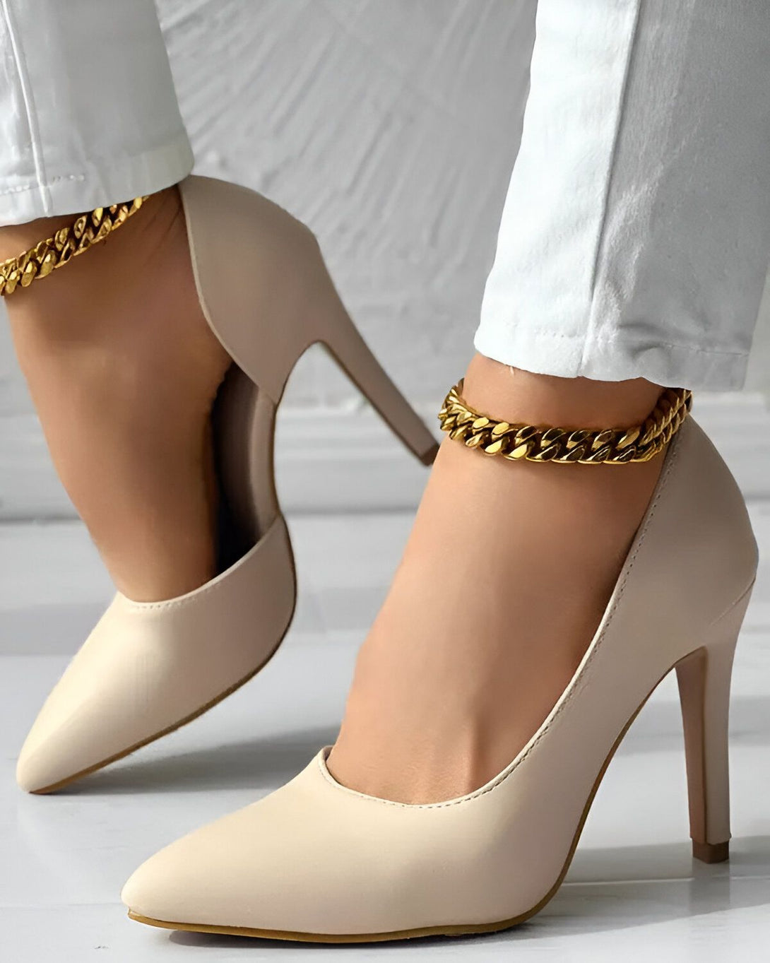 Une femme porte des escarpins beiges à talons hauts et fins. Les chaussures ont une chaîne dorée qui entoure la cheville. La femme porte un jean blanc et le bas des jambes est visible.