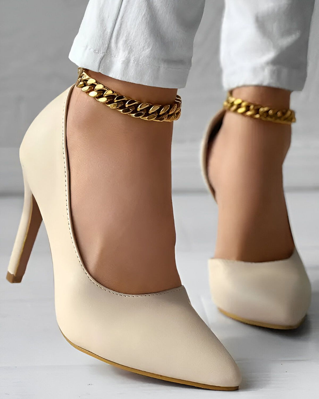 Une femme porte des escarpins blancs à talons hauts et des chaînettes dorées aux chevilles.