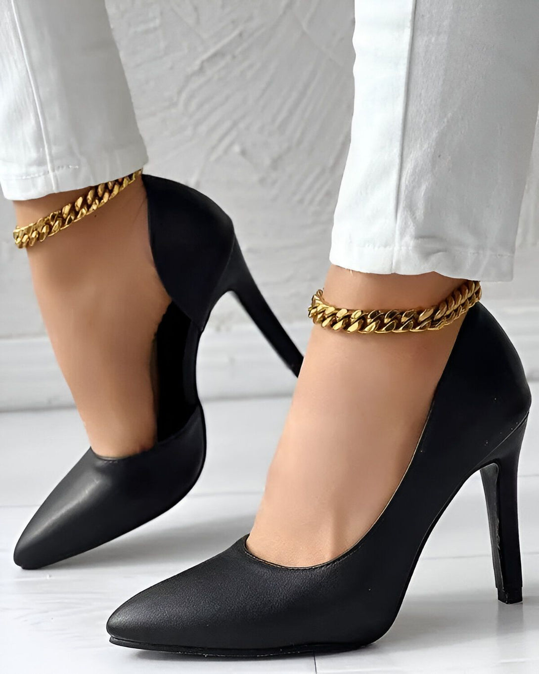 Une femme porte des escarpins noirs à talons hauts. Les chaussures ont une chaîne dorée autour de la cheville. La femme porte un jean blanc et les escarpins sont noirs.