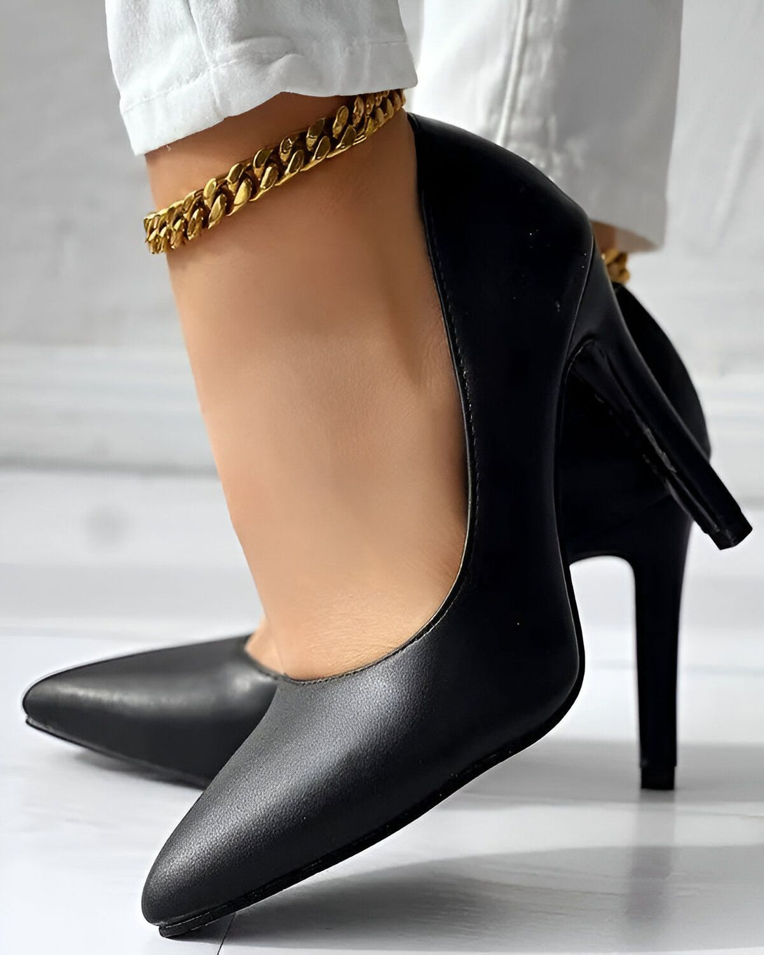 Une femme porte des escarpins noirs à talons hauts. Elle porte également un bracelet de cheville en or.