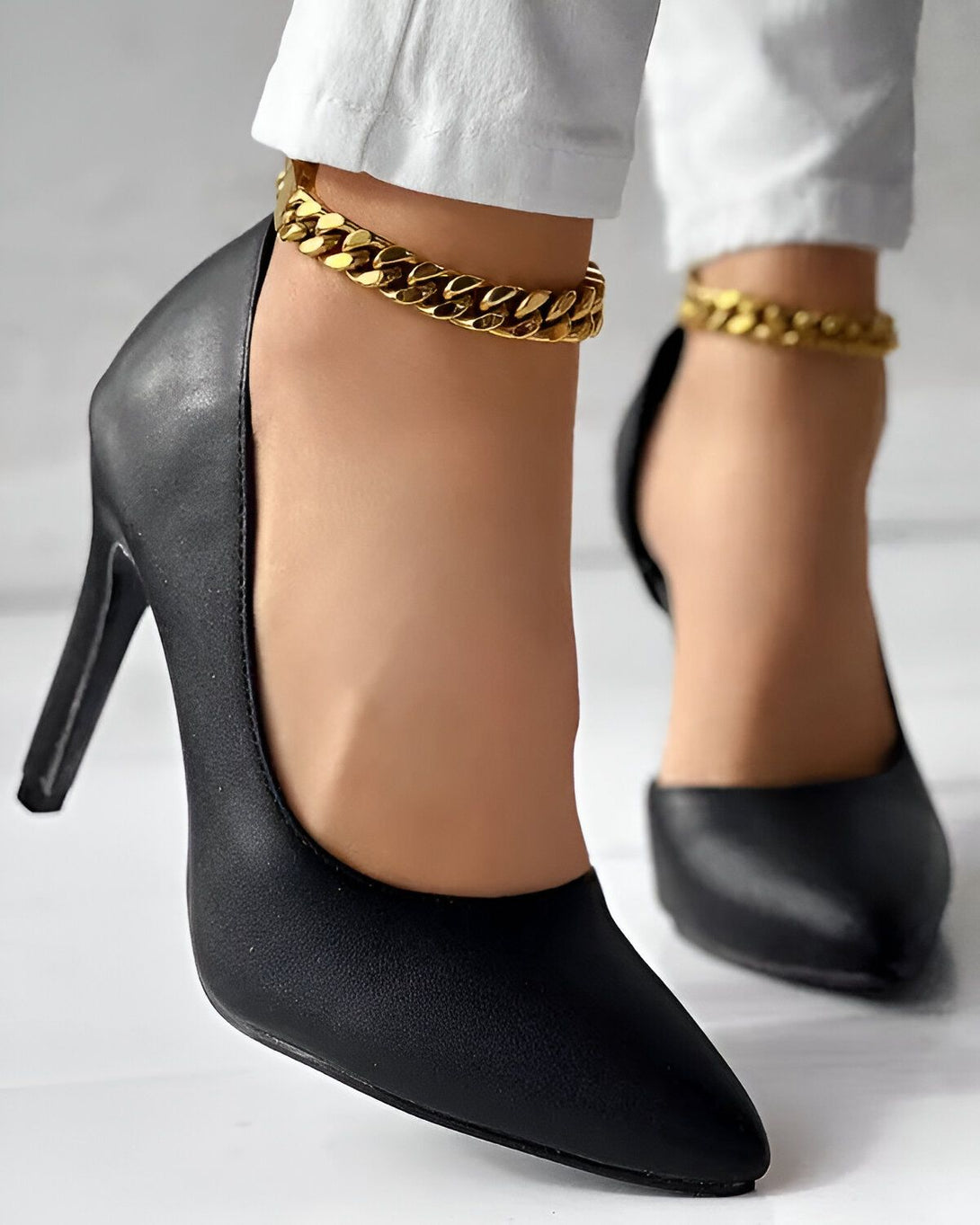 Une femme porte des escarpins noirs à talons hauts. Les chaussures ont une chaîne dorée autour de la cheville. La femme porte un jean blanc.