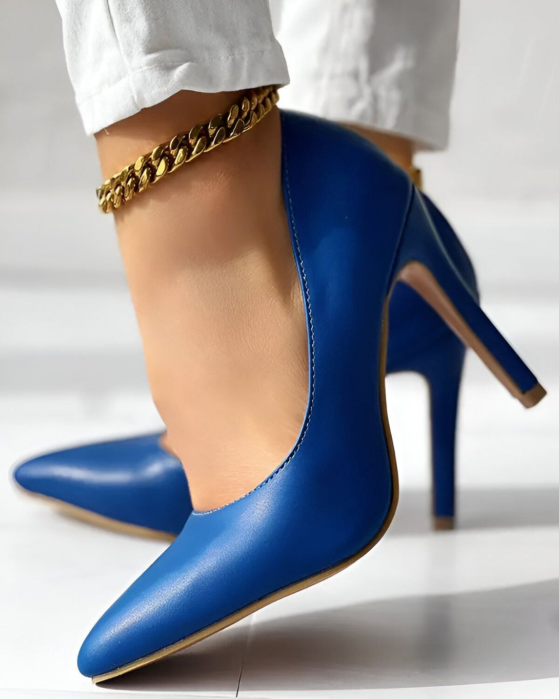 Une femme porte des escarpins bleus à talons hauts. Elle porte également un pantalon blanc et une cheville dorée. Les chaussures sont en cuir et ont un bout pointu.