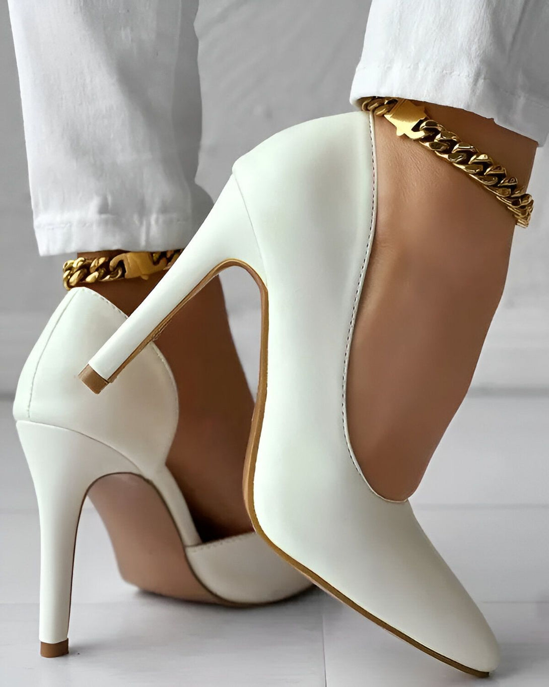 Une femme porte des escarpins blancs à talons hauts et un bracelet de cheville en chaîne dorée. Elle porte également un jean blanc.