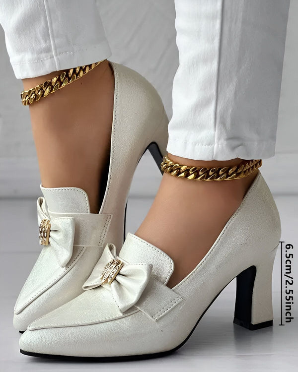 Une image d'une paire d'escarpins blancs à talons hauts avec un nœud sur le devant. Les escarpins sont portés par une femme qui porte un jean blanc et une chaîne dorée autour de la cheville.