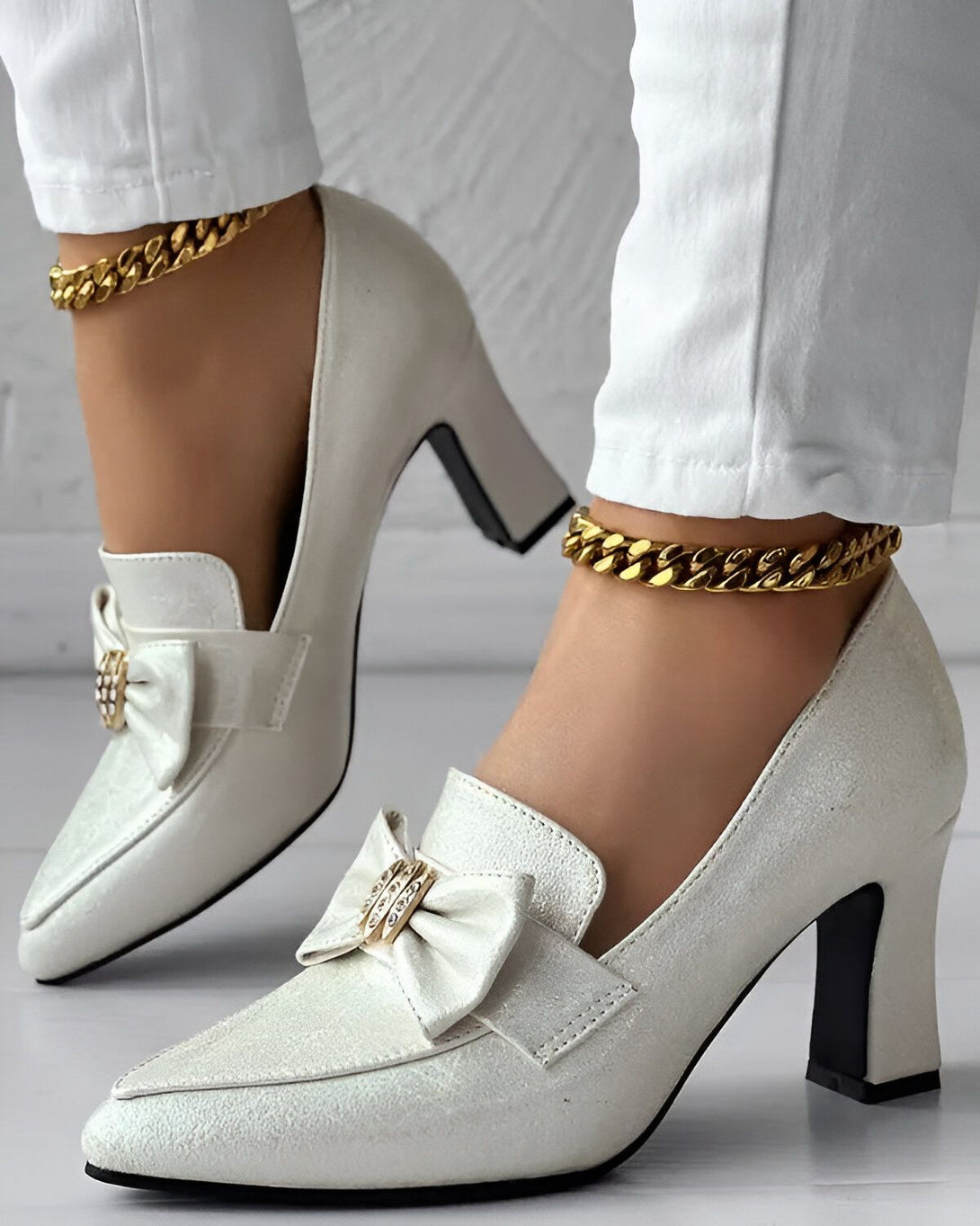 Une femme porte des escarpins blancs à talons hauts. Les escarpins sont ornés d'un nœud sur le devant. La femme porte également une chaîne dorée autour de la cheville.
