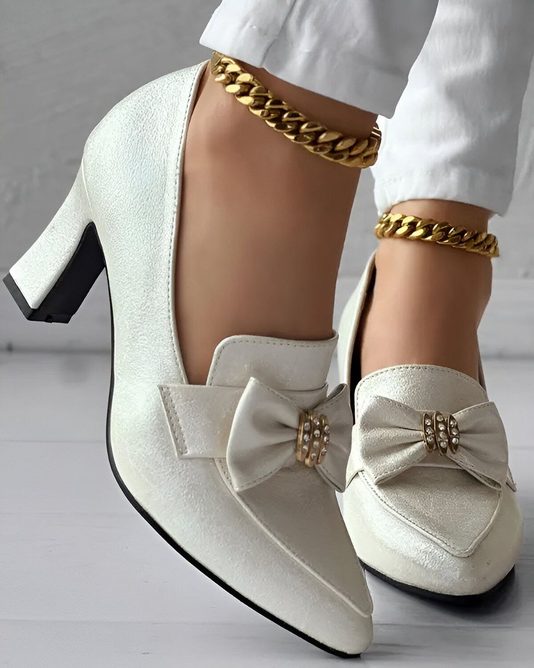 Une paire d'escarpins blancs à talons hauts avec un nœud en satin sur le devant. Les chaussures sont portées par une femme qui porte également un pantalon blanc et des bijoux en or.
