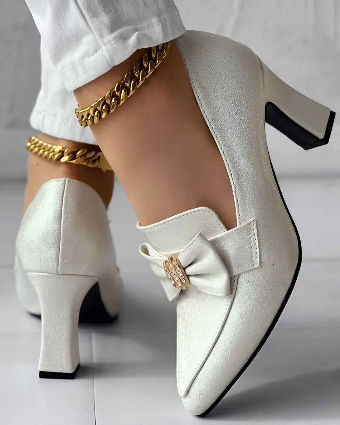 Une femme porte des escarpins blancs à talons hauts. Les chaussures sont en cuir verni et ont un nœud sur le devant. La femme porte également un pantalon blanc et un bracelet de cheville en or.