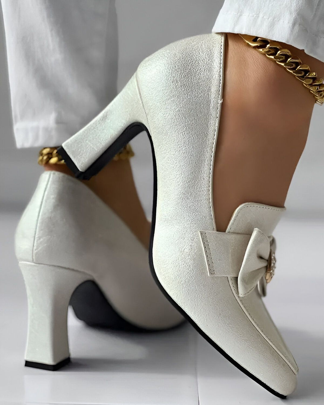 Une paire d'escarpins blancs à talons hauts avec une boucle et un nœud en strass. Les chaussures sont portées par une femme qui porte un pantalon blanc et une cheville dorée.