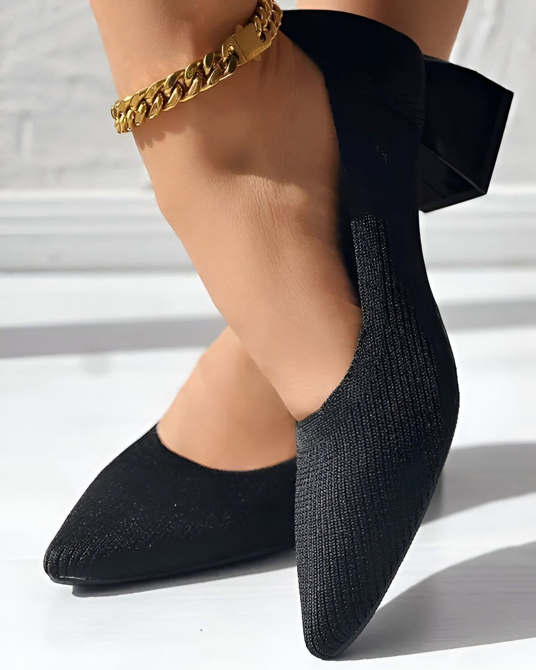 Une femme porte des chaussures noires à talons et un bracelet de cheville en or. Les chaussures sont en maille et ont un bout pointu. Le talon est épais et mesure environ 5 cm. La femme est debout sur un sol en marbre blanc.