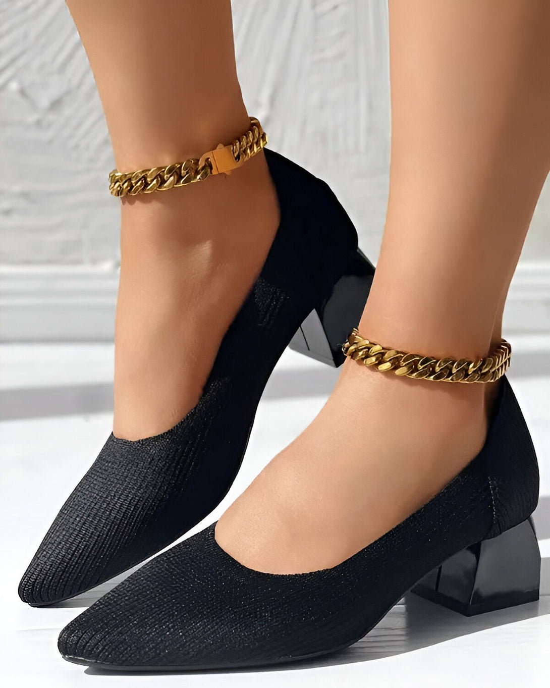 Une femme porte des escarpins noirs à talons hauts avec des chaînes dorées autour des chevilles.
