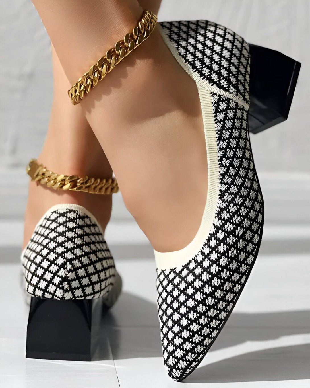 Une femme porte des chaussures à talons blancs et noirs avec un motif en losange. Les chaussures ont un bout pointu et un talon épais. La femme porte également une chaîne dorée autour de la cheville.