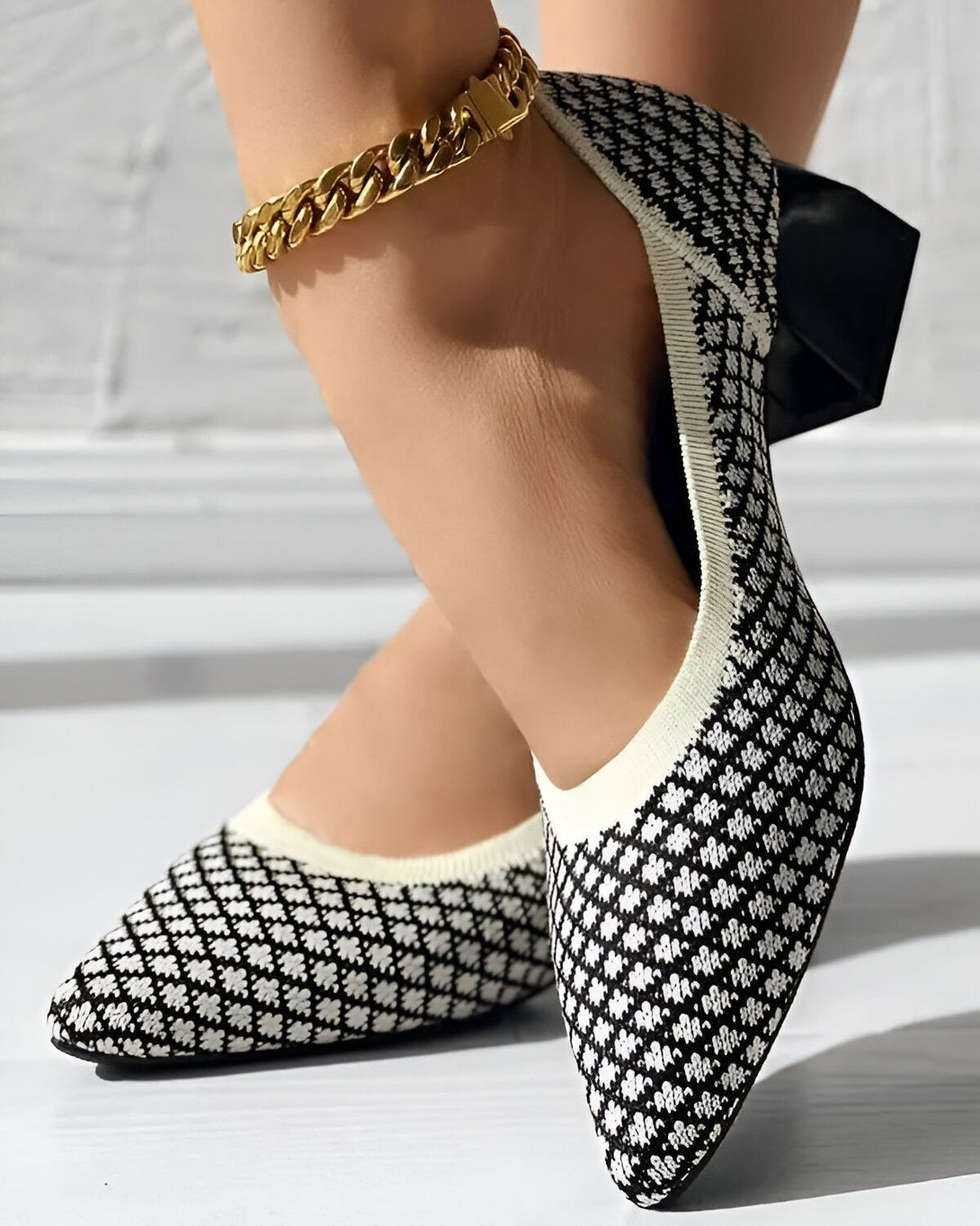 Une femme porte des chaussures noires et blanches à talons. Les chaussures sont en maille et ont un motif de losanges. La femme a une chaîne dorée à la cheville.