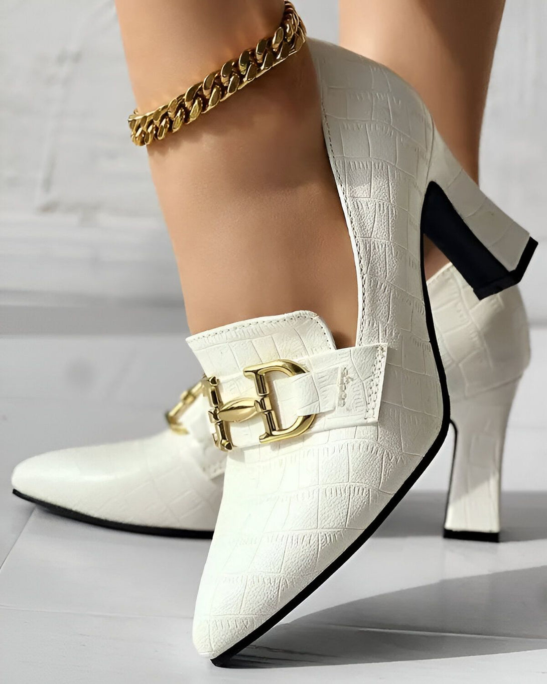 Une image d'une paire d'escarpins blancs à talons hauts avec une boucle dorée sur le devant. Les escarpins sont portés par une femme qui a une chaîne dorée autour de la cheville.