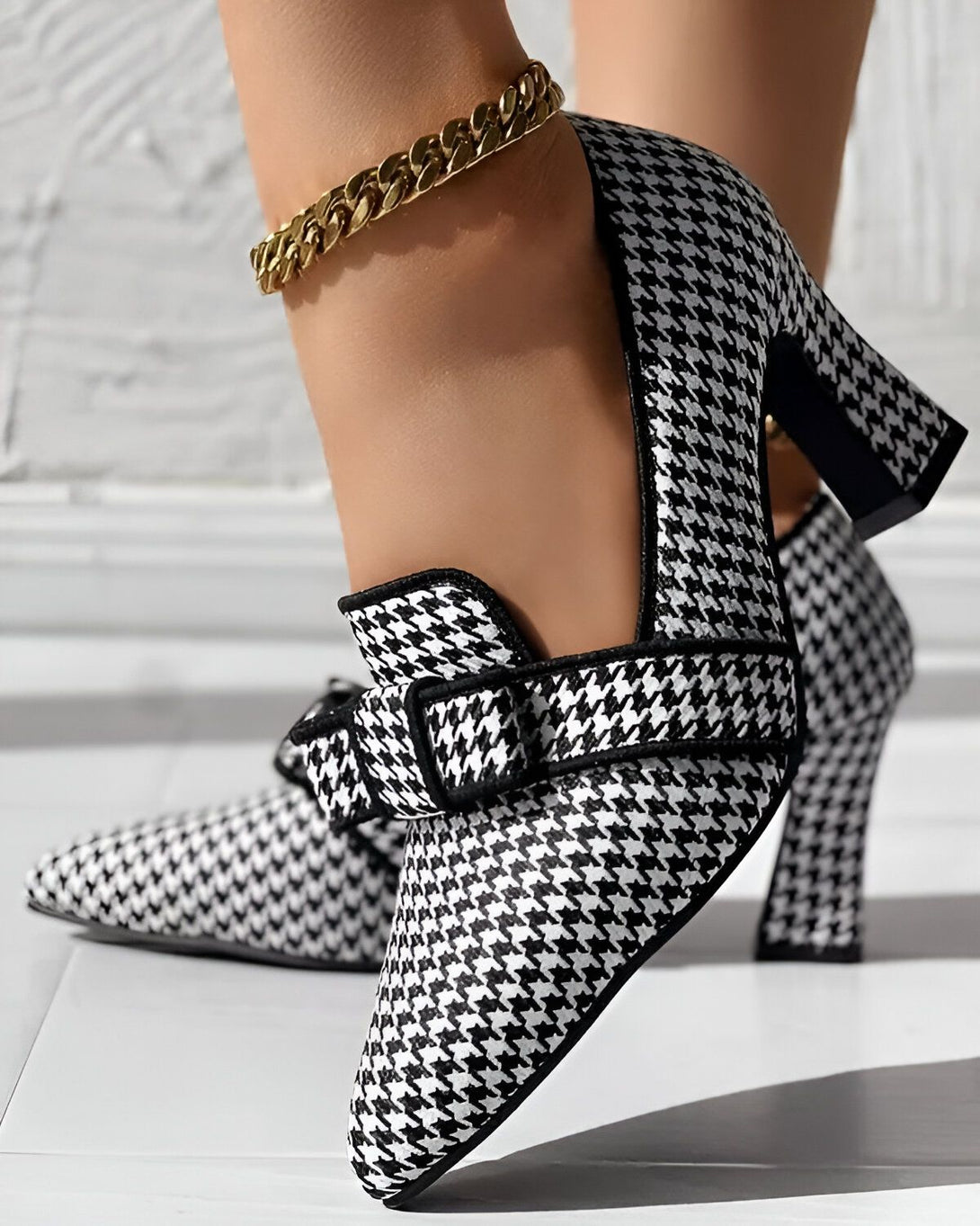 Une image d'une paire d'escarpins à talons hauts noirs et blancs à motif pied-de-poule. Les chaussures sont portées par une femme qui porte également une chaîne de cheville en or.