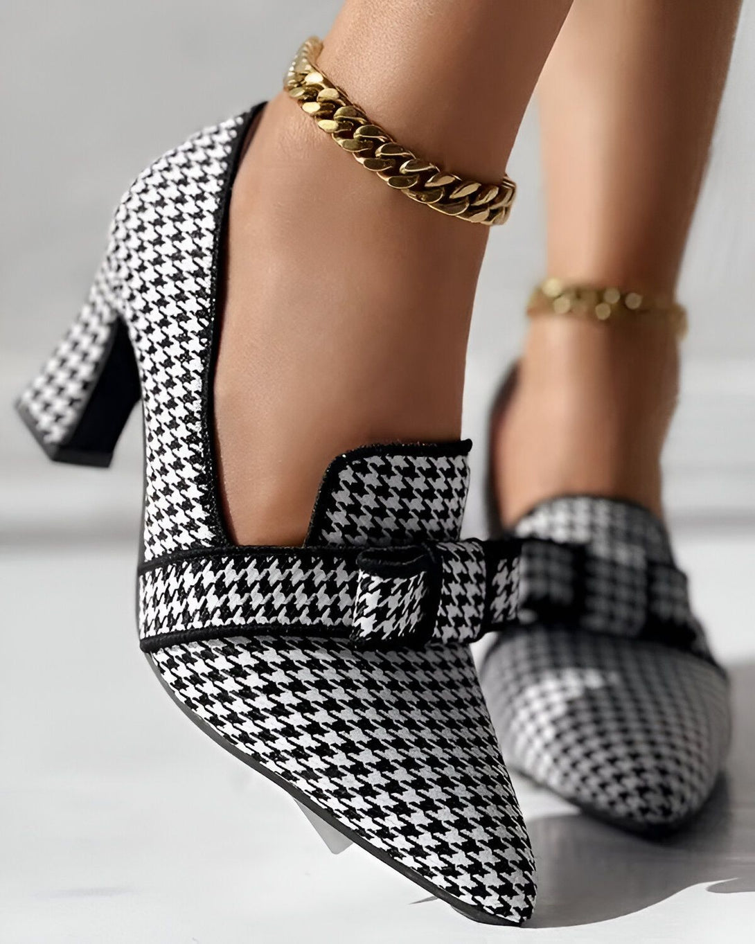 Une image d'une paire d'escarpins à talons hauts noirs et blancs à motif pied-de-poule. Les escarpins sont portés par une femme qui porte également une chaîne dorée autour de la cheville.