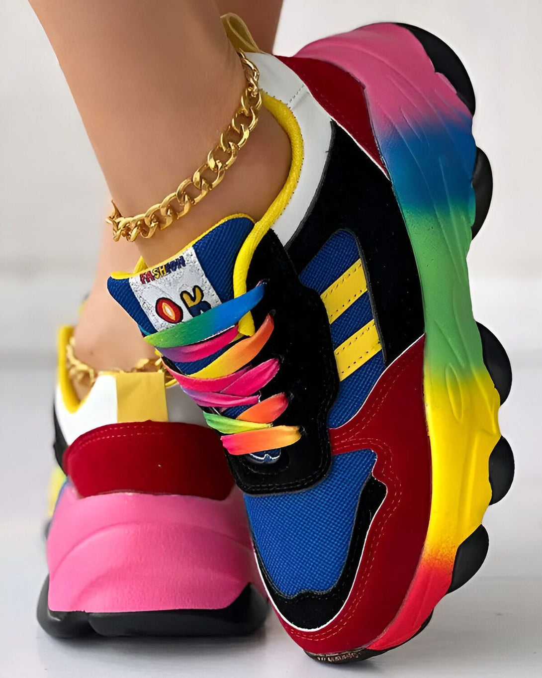 Une paire de baskets multicolores avec des lacets arc-en-ciel sur un pied et une chaîne de cheville dorée sur l'autre.