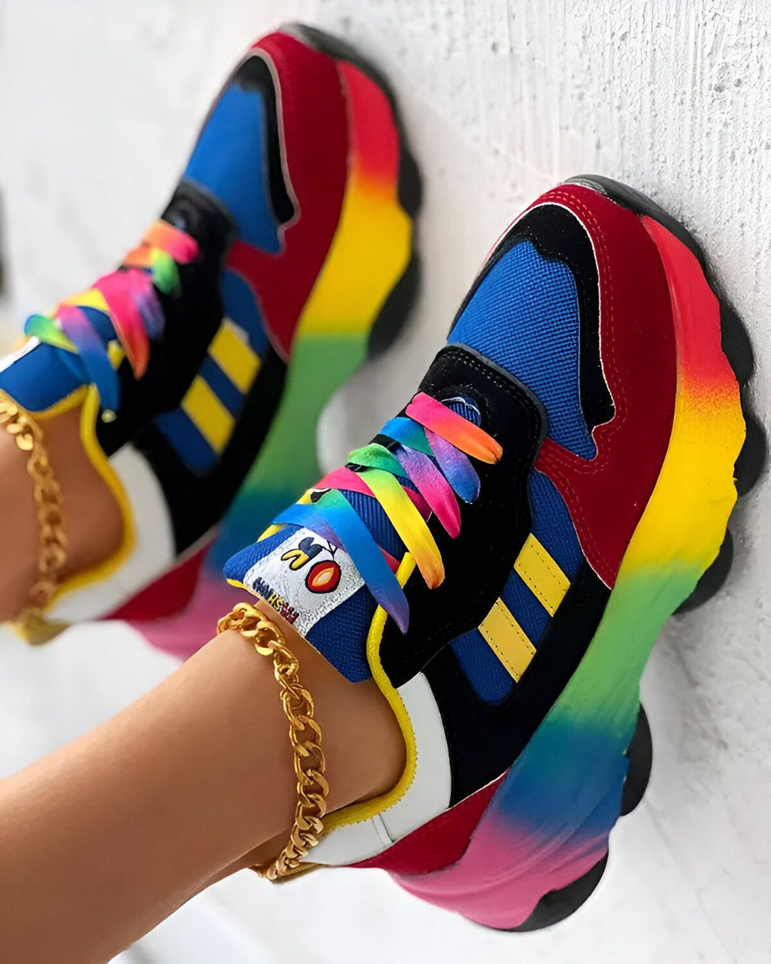 Une paire de baskets multicolores avec des lacets arc-en-ciel sur un pied avec une cheville avec une chaîne en or.