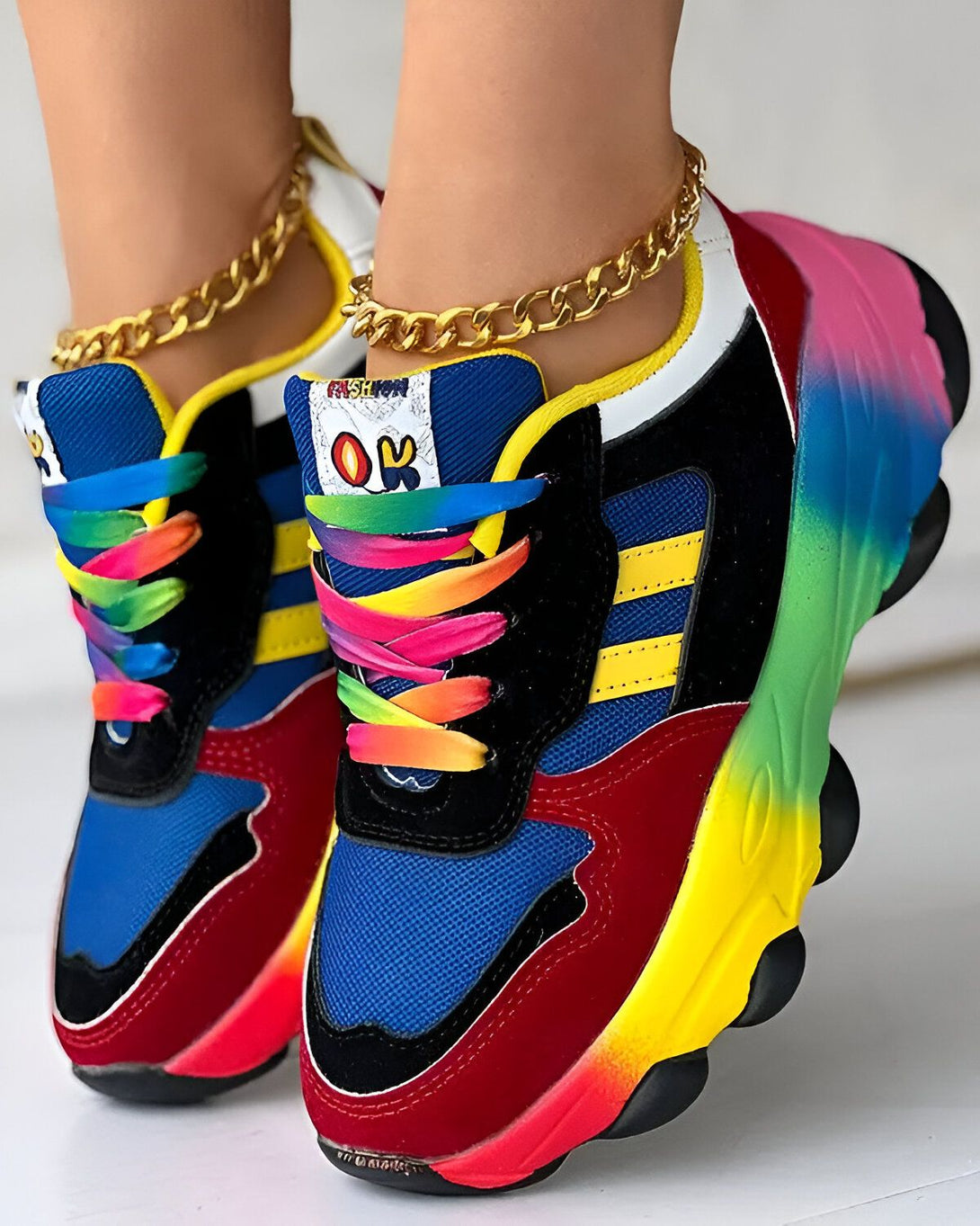 Une paire de baskets multicolores avec des lacets arc-en-ciel sur un pied et une chaîne dorée sur l'autre.