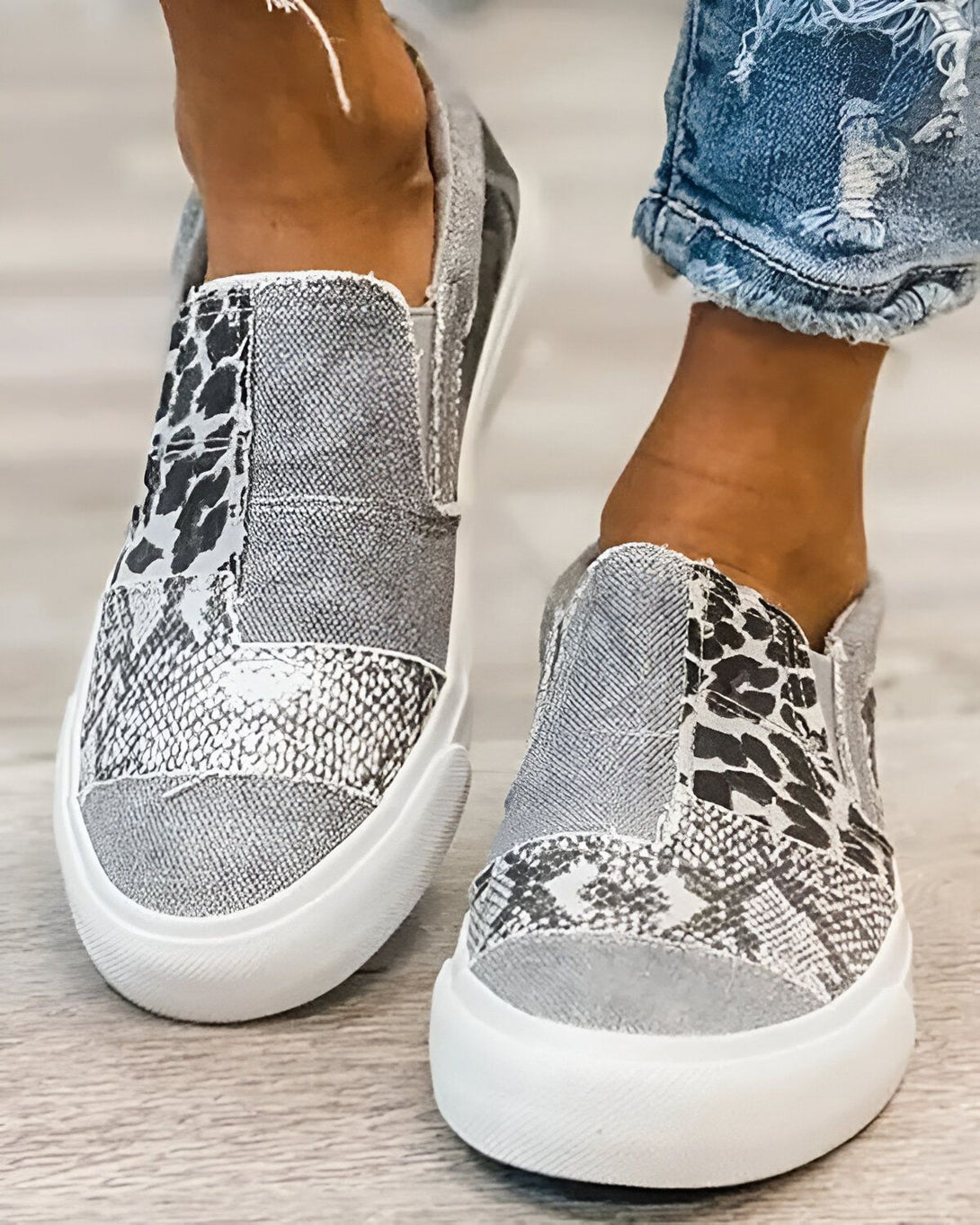 Une femme porte des baskets grises et blanches. Les baskets sont en patchwork de différents matériaux, notamment du daim, du cuir et du textile. La femme porte un jean bleu troué.