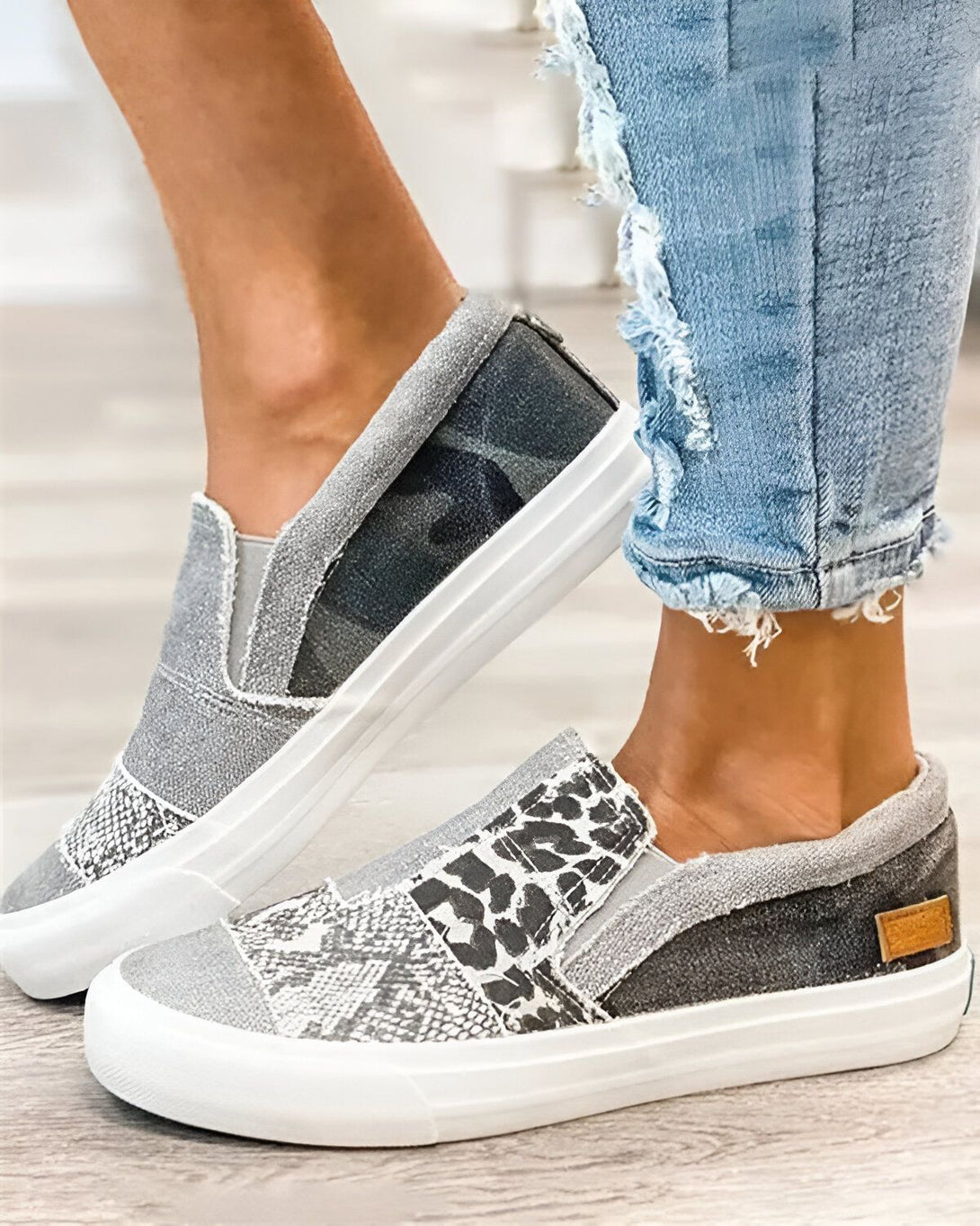Une femme porte des chaussures slip-on grises avec des motifs léopard et camouflage. Elle porte également un jean bleu déchiré.