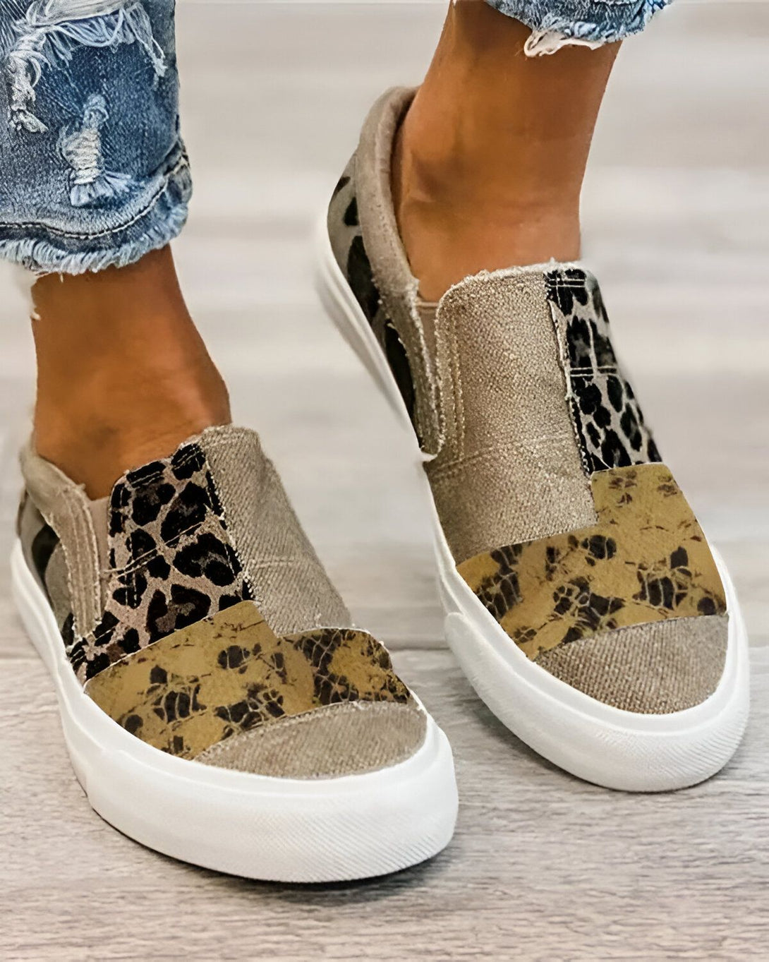 Une femme porte des chaussures slip-on en toile beige avec des empiècements en simili cuir marron et noir. Les chaussures ont une semelle blanche.
