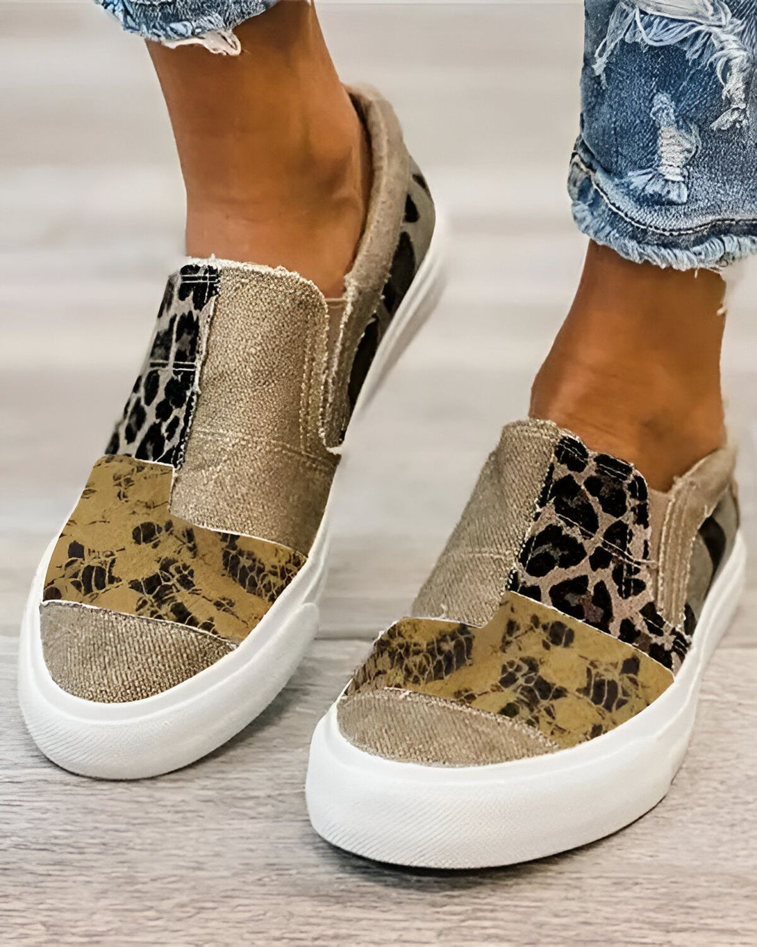 Une femme porte des chaussures slip-on beiges et marron avec un motif léopard. Les chaussures ont une semelle blanche. La femme porte un jean bleu troué.