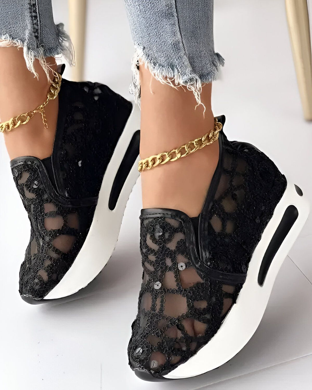 Une femme porte des baskets noires en dentelle avec une semelle blanche. Elle porte également un jean bleu déchiré et une cheville dorée.