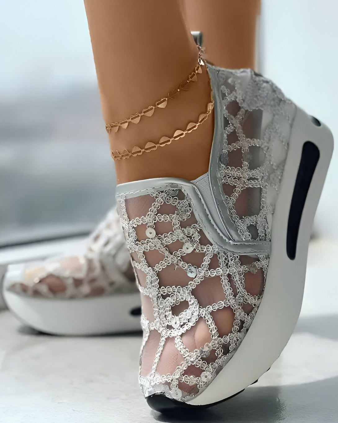 Une femme porte des baskets blanches en dentelle avec des paillettes argentées. Les baskets sont lacées et la femme porte des bracelets de cheville en or.