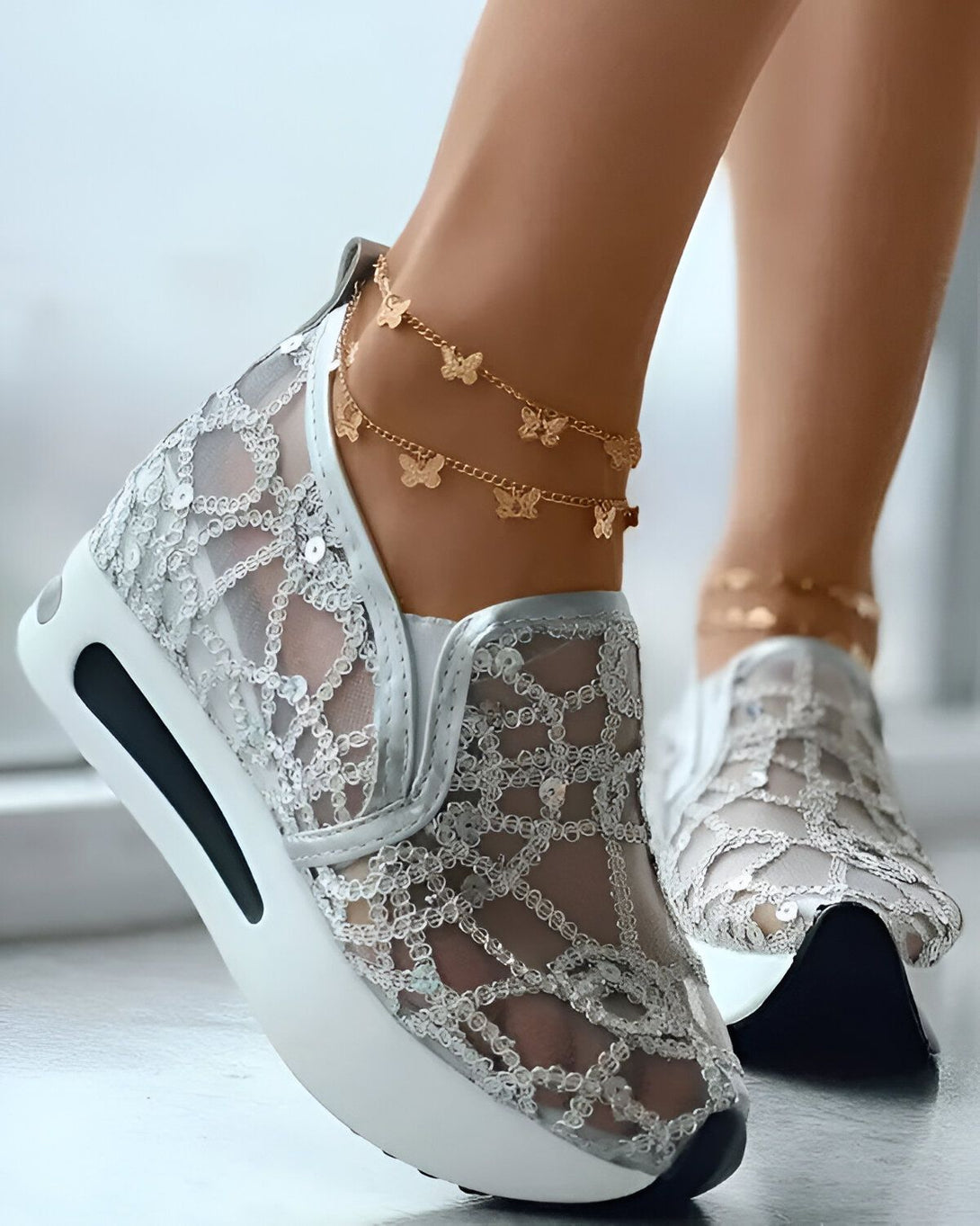 Une femme porte des baskets blanches à lacets avec un motif de papillons argentés. Elle porte également des bracelets de cheville dorés avec des pendentifs papillons.