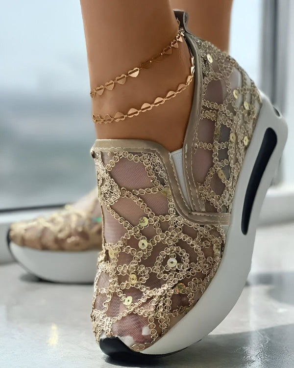 Une femme porte des baskets dorées à lacets avec des paillettes et un filet transparent. Les baskets ont une semelle blanche et une bride velcro. La femme porte également des bracelets de cheville en or.