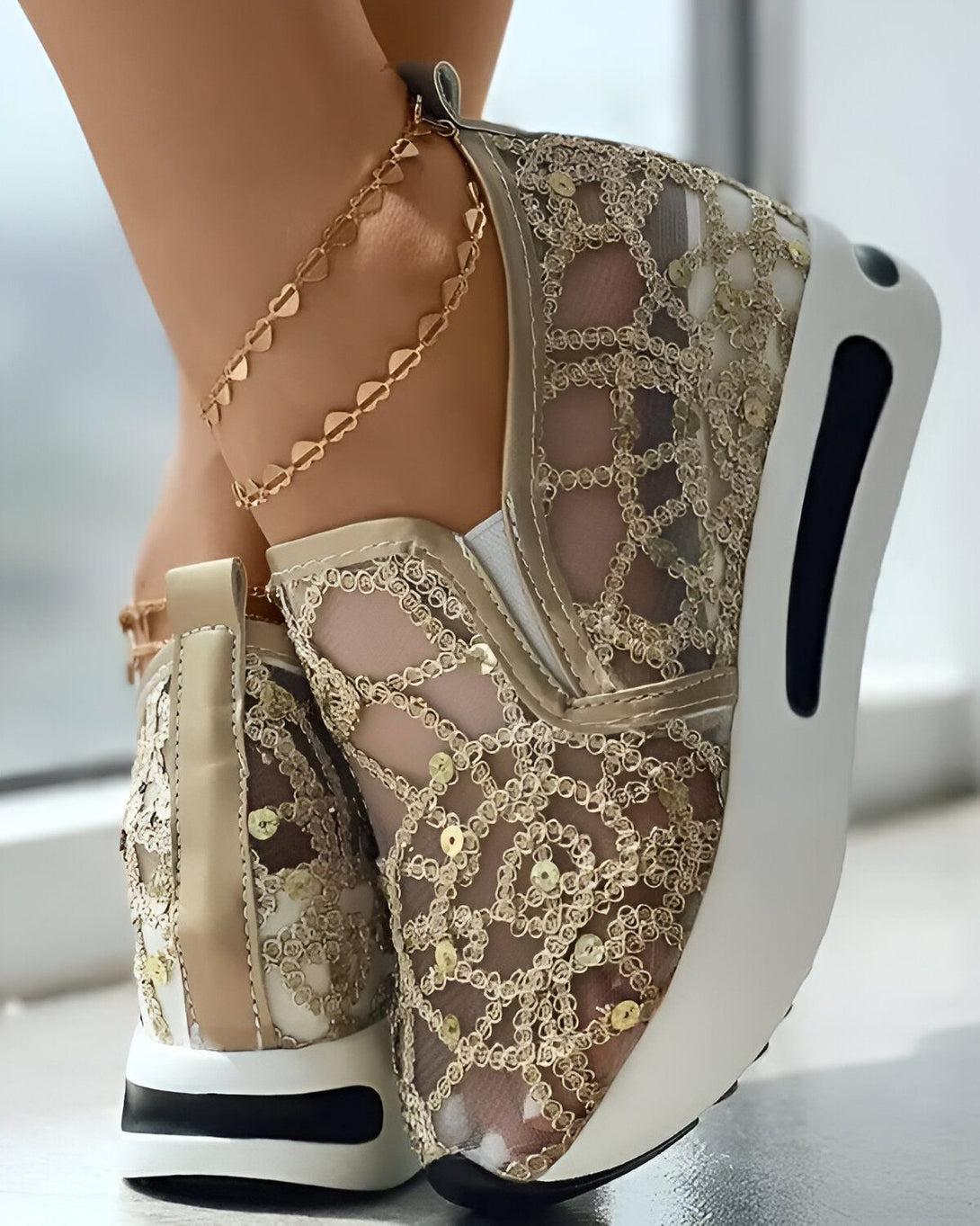 Une femme porte des baskets dorées en dentelle avec des paillettes. Les baskets ont une semelle blanche et sont lacées avec des lacets dorés. La femme porte également une chaîne de cheville en or.