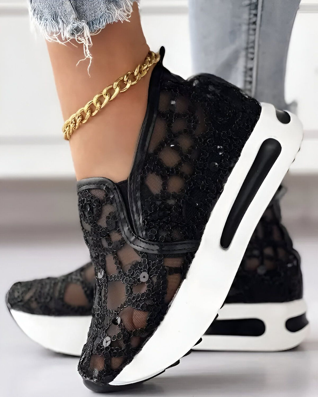 Une femme porte des baskets noires en dentelle avec des paillettes. Les baskets ont une semelle blanche et des lacets noirs. La femme porte également un jean bleu et un chemisier blanc.