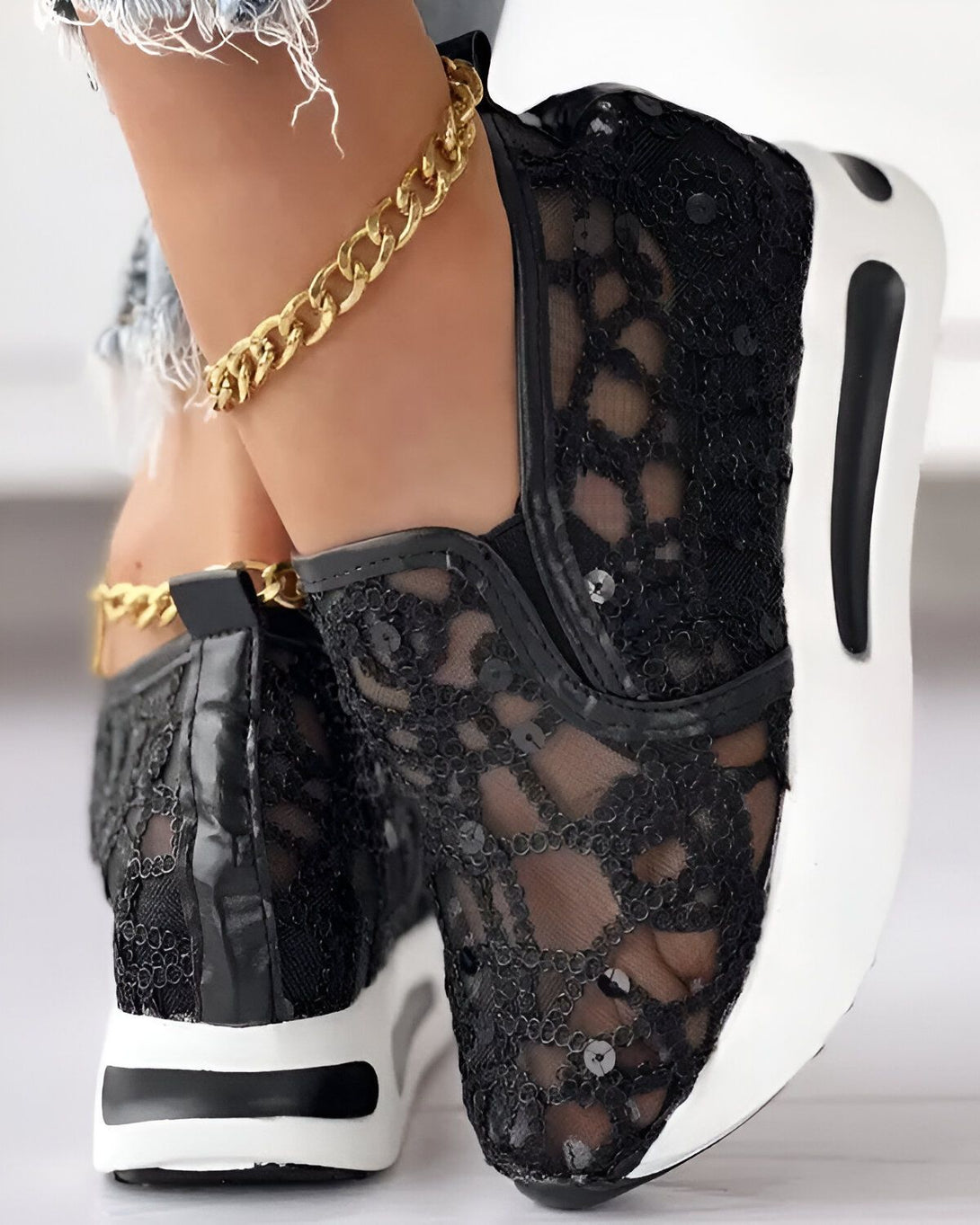 Une femme porte des baskets noires en dentelle avec des paillettes. Les baskets sont lacées et ont une semelle blanche. La femme porte également un jean bleu et un bracelet de cheville en or.