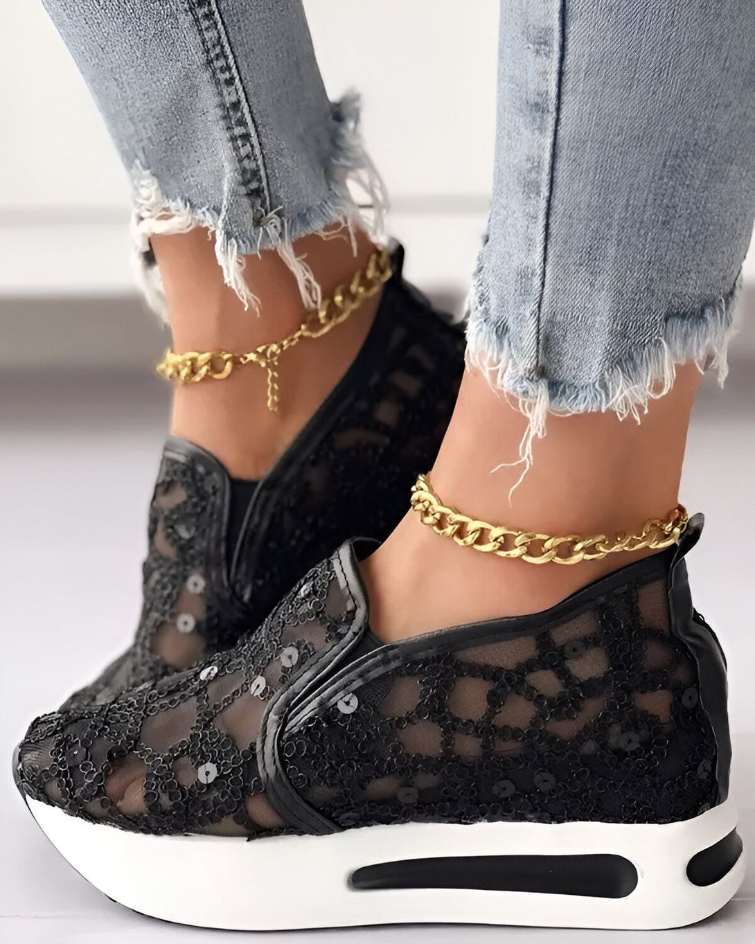 Une femme porte des baskets noires en dentelle avec des semelles blanches. Elle porte également un jean bleu déchiré et des bracelets de cheville en or.