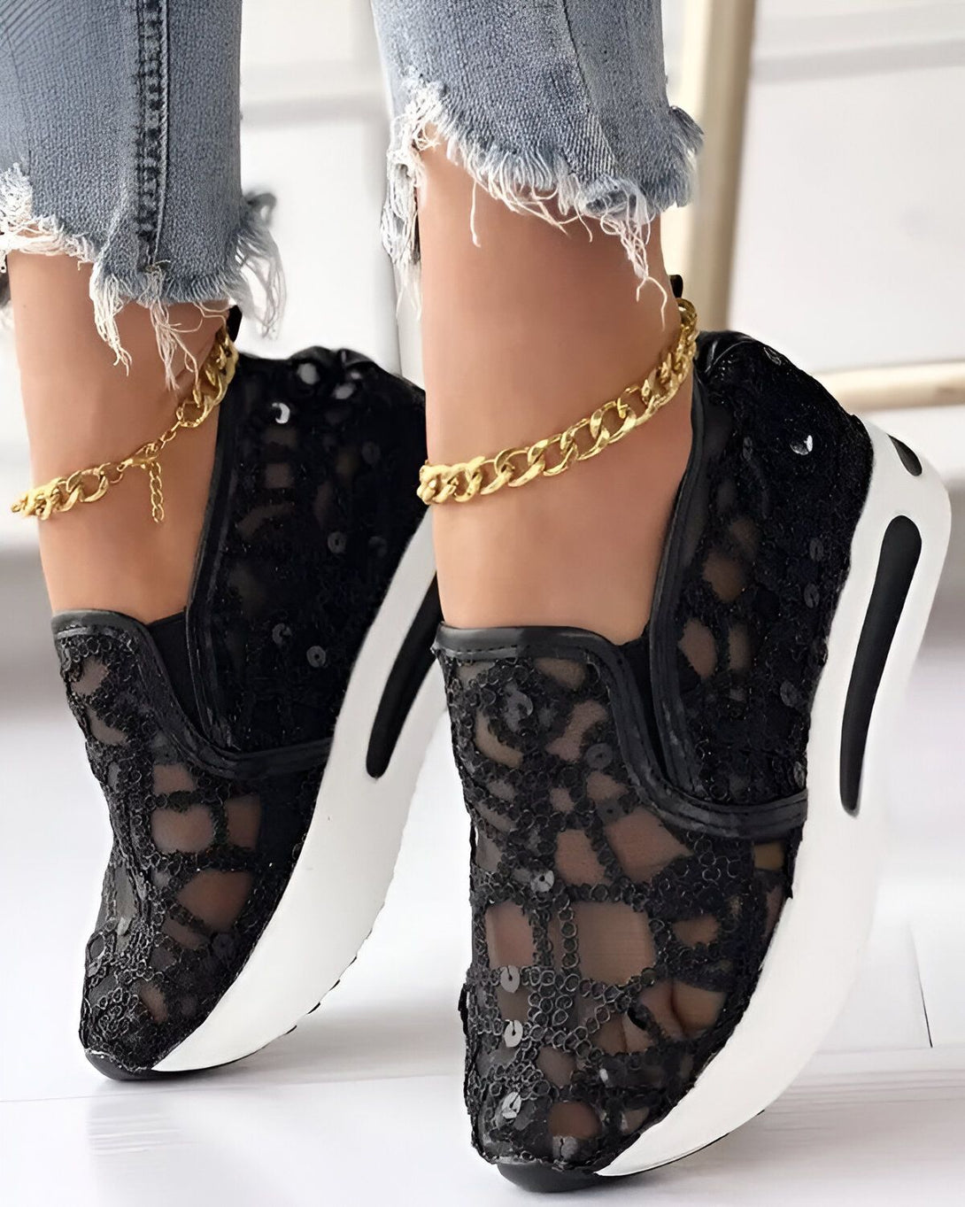Une femme porte des baskets noires en dentelle avec des paillettes. Les baskets ont une semelle blanche et des lacets noirs. La femme porte également un jean bleu et un bracelet de cheville en or.