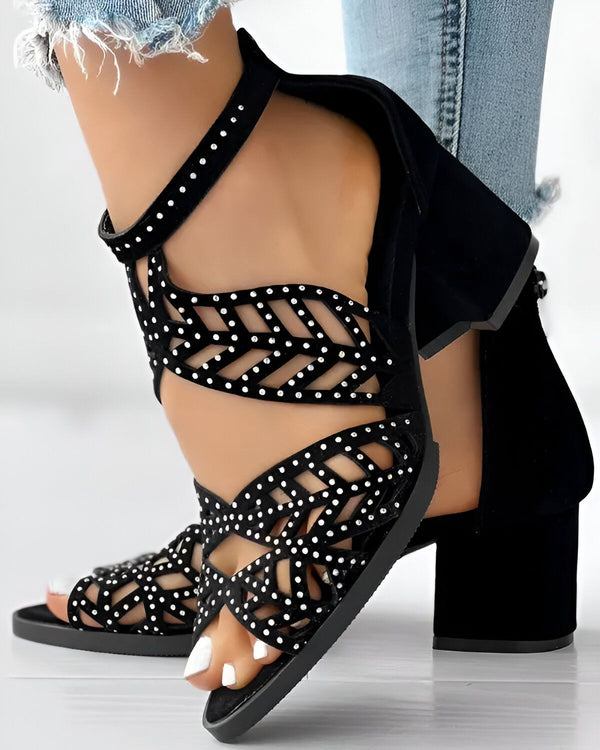 Une femme porte des sandales noires à talons hauts. Les sandales sont ornées de strass et ont une bride qui s'enroule autour de la cheville. La femme porte un jean bleu et un haut blanc.