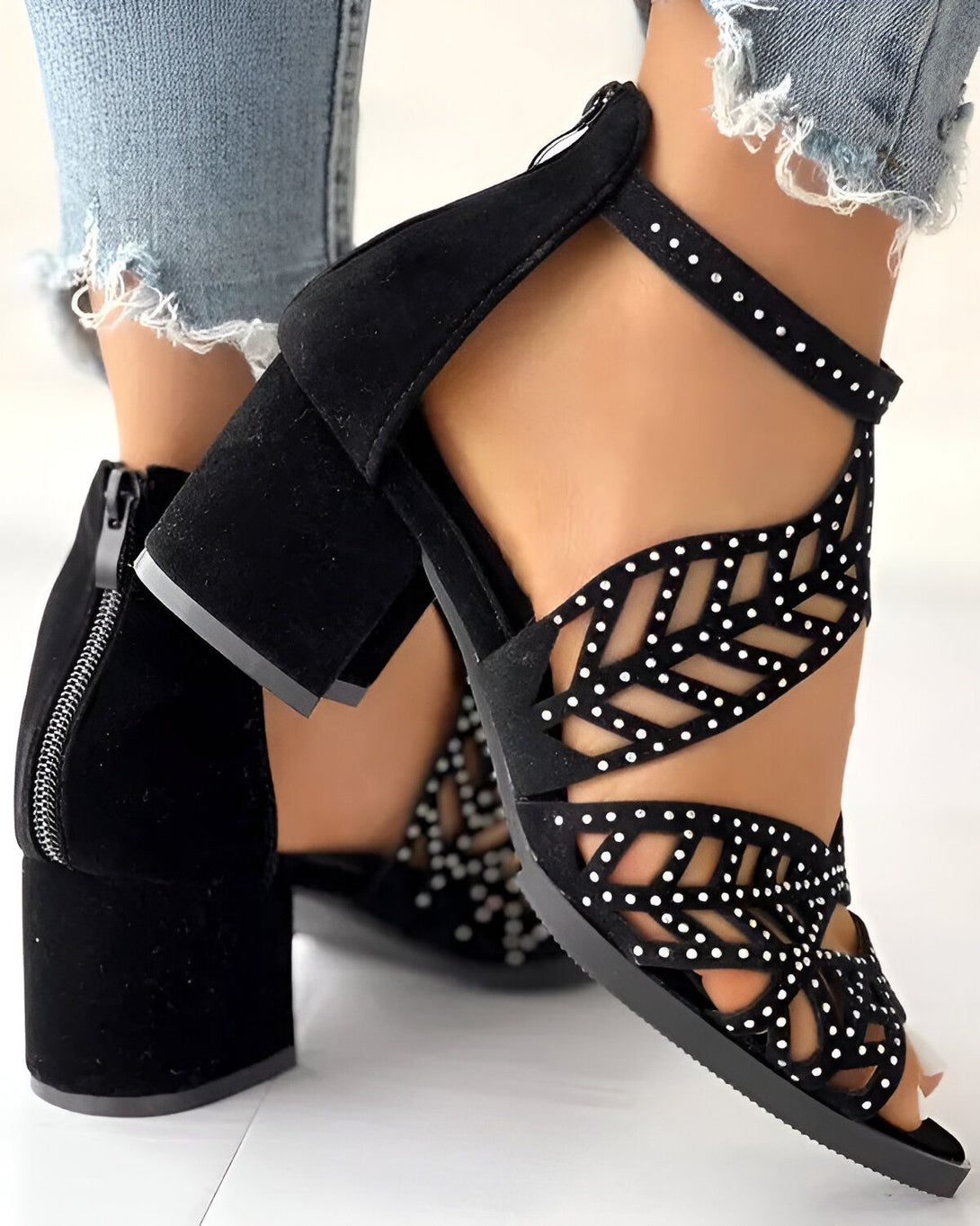 Une femme porte des sandales noires à talons hauts. Les sandales sont en daim et ont des détails cloutés. La femme porte un jean bleu déchiré.