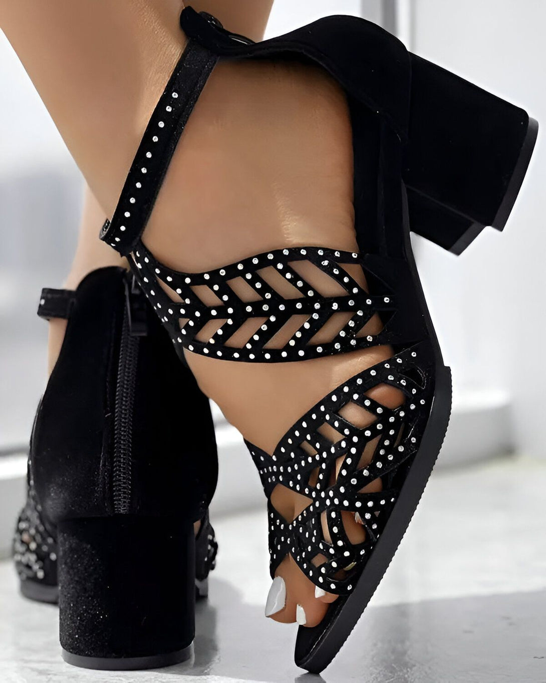 Une femme porte des sandales noires à talons hauts. Les sandales sont en daim et ont des brides croisées sur le devant. Le talon est épais et recouvert de strass. La femme a les ongles des pieds peints en blanc.