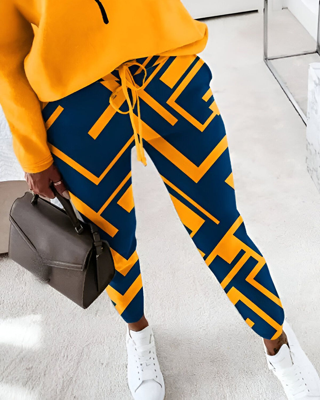 Une femme porte un pantalon de survêtement bleu et jaune avec des baskets blanches et un haut jaune. Elle porte également un sac à main marron.