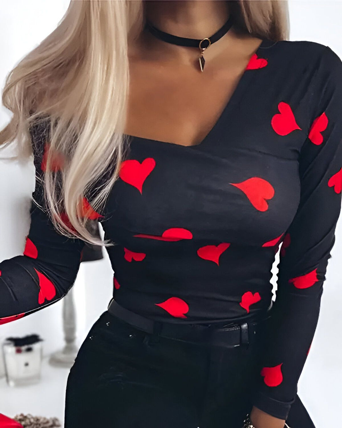 Une femme porte un haut moulant noir à manches longues avec un imprimé de cœurs rouges. Elle porte également un collier noir avec un pendentif en forme de cœur.