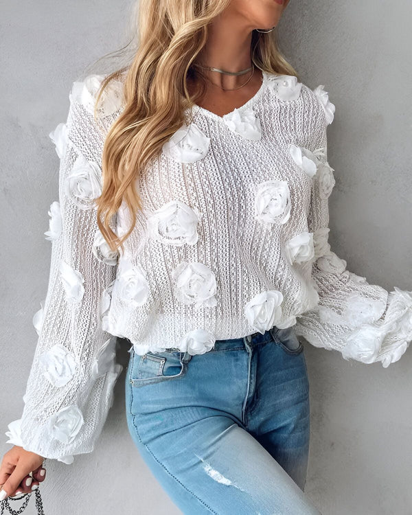 Une jeune femme porte un pull blanc avec des fleurs en relief. Elle est également vêtue d'un jean bleu et porte un collier.