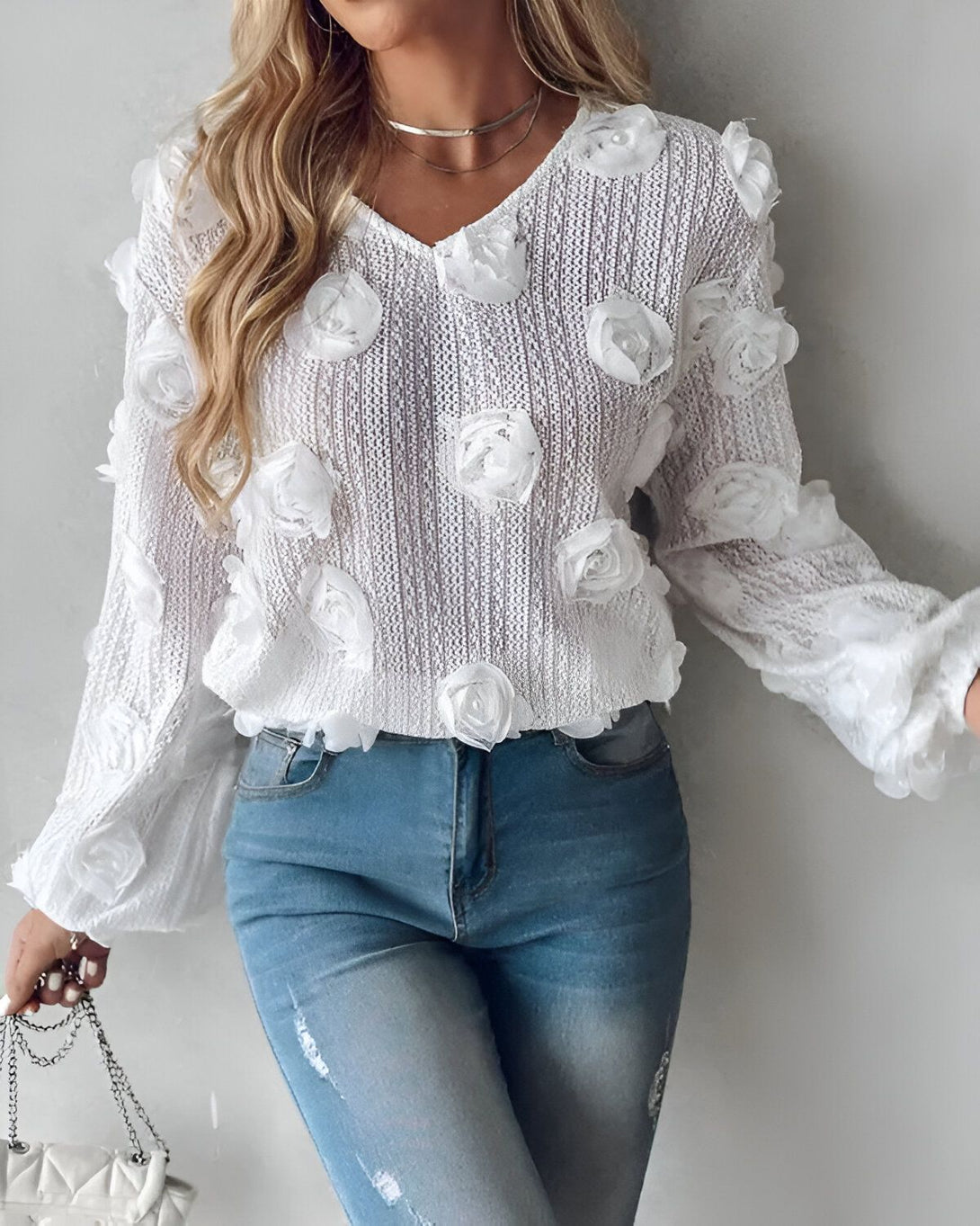 Une jeune femme porte un pull blanc avec des fleurs en relief, un jean bleu et un collier argenté.