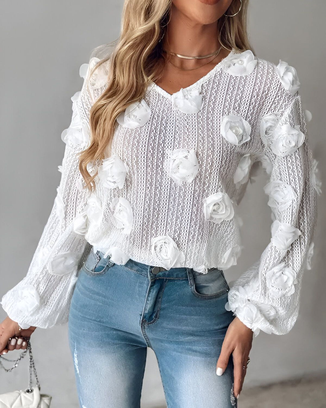 Une jeune femme porte un pull blanc avec des fleurs en relief. Le pull est en laine et a des manches longues. La femme porte également un jean bleu et des talons hauts blancs.