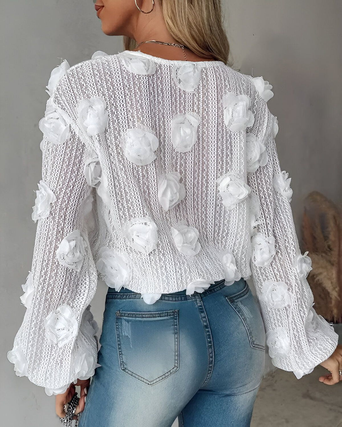 Une femme porte un pull blanc avec des fleurs en relief. Le pull est court et les manches sont bouffantes. La femme est vue de dos. Elle porte un jean bleu et des talons hauts.