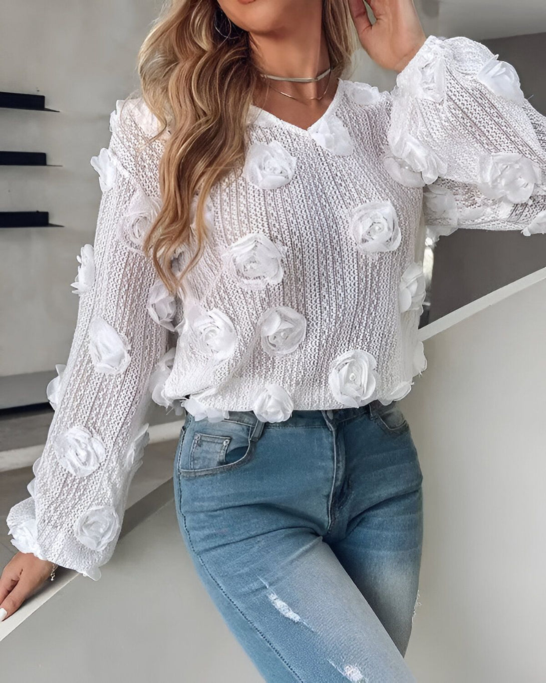 Une femme porte un pull blanc avec des fleurs en relief et un jean bleu troué.