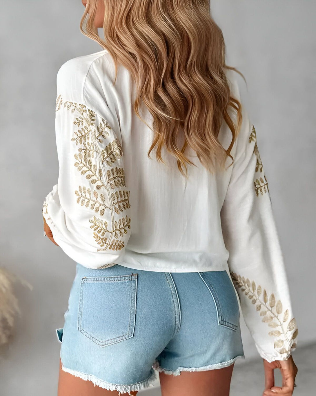Une femme porte un chemisier blanc avec des broderies dorées et un short en jean bleu.