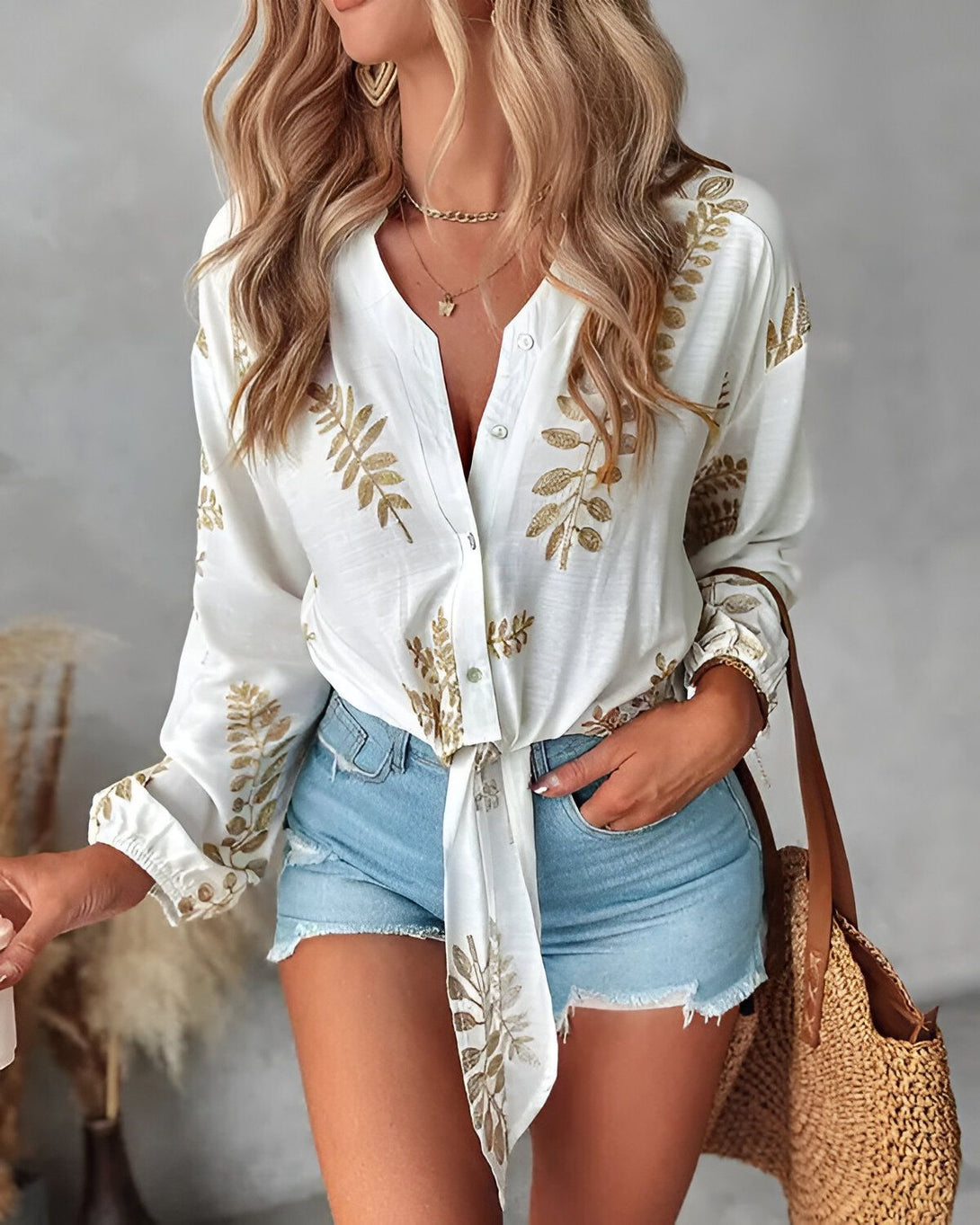 Une femme porte une blouse blanche à motifs de feuilles dorées, un short en jean bleu et un sac à main marron.