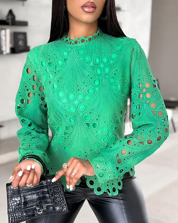 Une femme porte un chemisier vert à col montant et à manches longues. Le chemisier est ajouré et présente un motif de cercles. La femme porte également un pantalon en cuir noir et un sac à main noir.