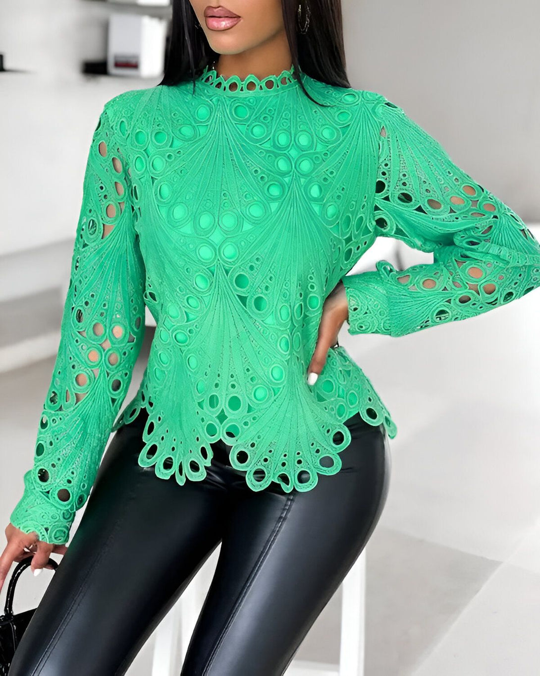 Une femme porte un chemisier vert à col montant et à manches longues. Le chemisier est ajouré et présente un motif de cercles. La femme porte également un pantalon en cuir noir et des talons noirs.