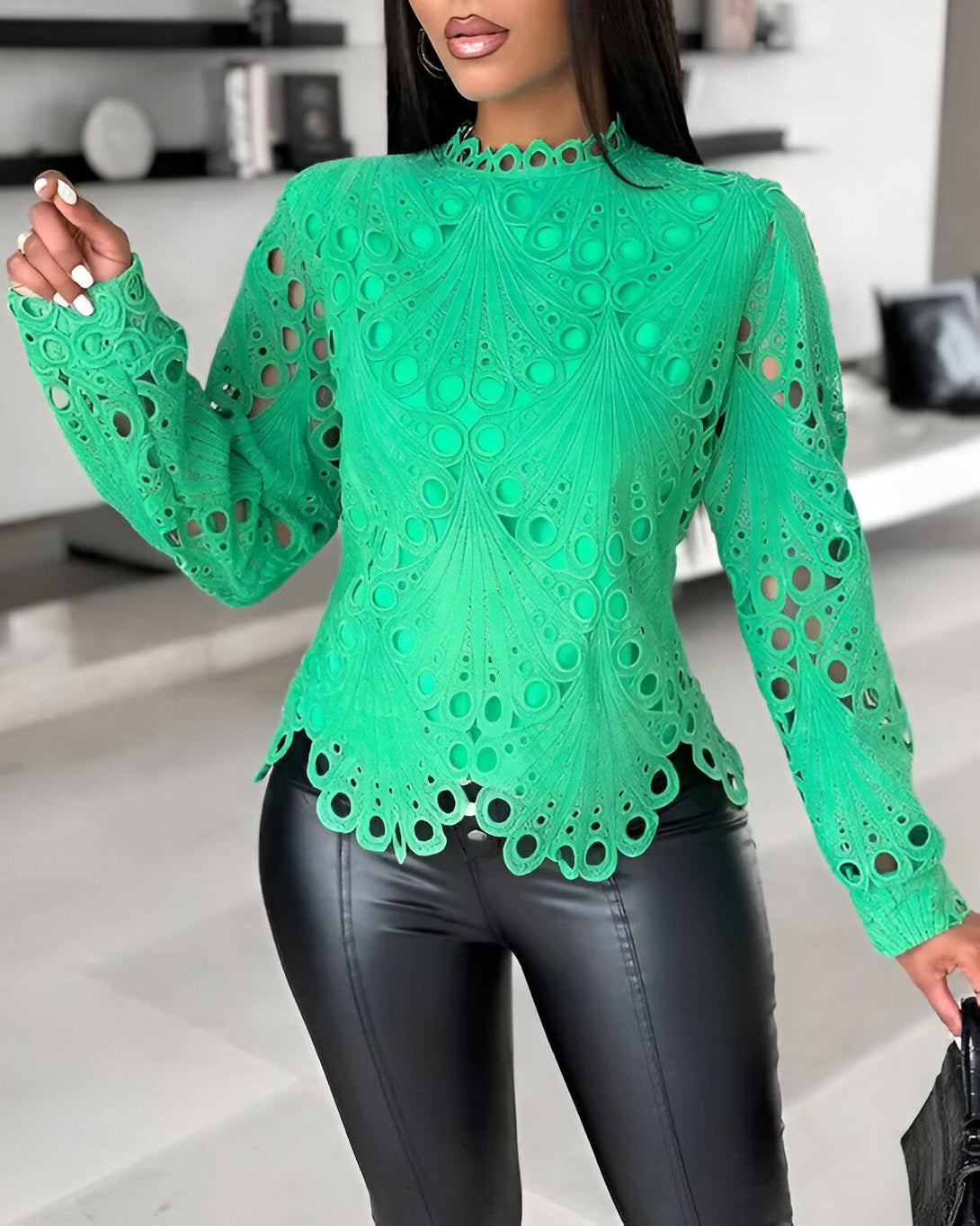 Une femme porte un chemisier vert à manches longues. Le chemisier est en dentelle et présente un col montant. La femme porte également un pantalon en cuir noir et des talons noirs.